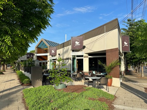 A modern restaurant exterior with outdoor seating, large windows, and greenery on a sunny day.