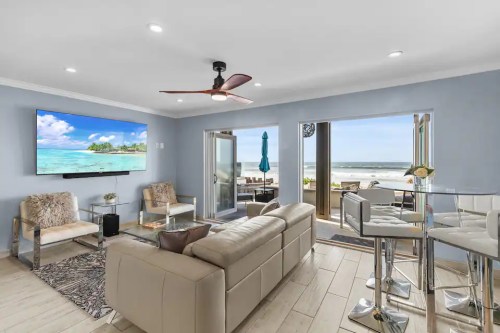 Modern beachside living room with a large TV, light-colored furniture, and ocean views through sliding glass doors.