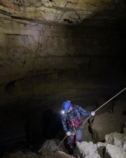 A person in a helmet and headlamp descends into a dark cave, using a rope for support on rocky terrain.