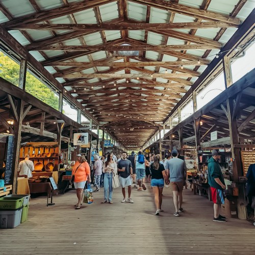 A bustling market with wooden beams, people walking between vendor stalls, and bright displays of goods.