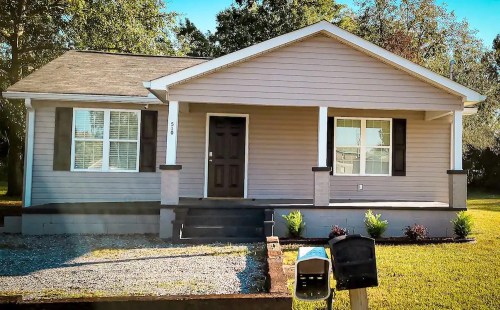 A single-story, light brown house with a front porch, two windows, and a landscaped yard. Mailboxes are visible in front.