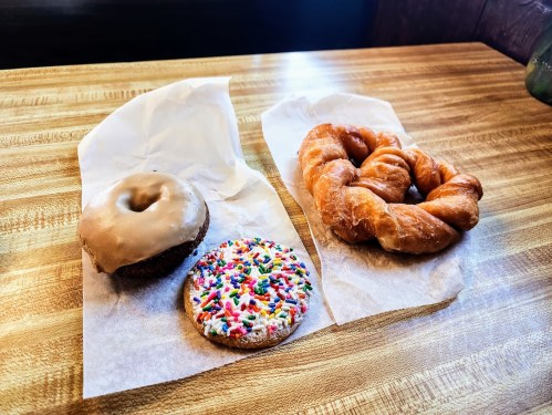 A glazed donut, a cookie with sprinkles, and a croissant on parchment paper on a wooden table.