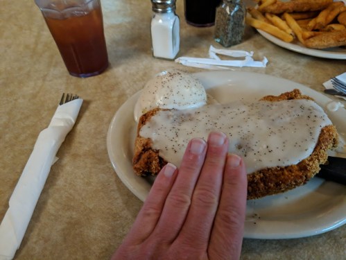 A hand rests on a plate with fried chicken covered in gravy, alongside a side of mashed potatoes and a drink in the background.