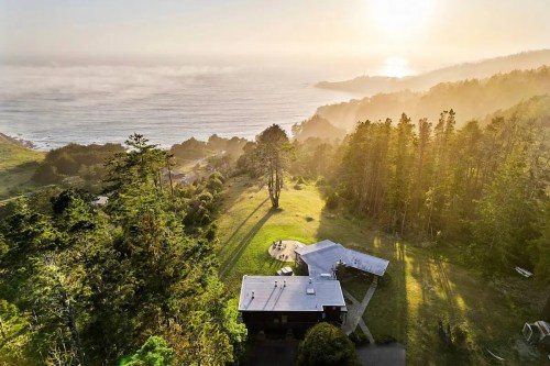 Aerial view of a coastal landscape with a modern house, surrounded by trees and sunlight illuminating the ocean.