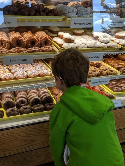 A child in a green hoodie gazes at a display of various donuts in a bakery.