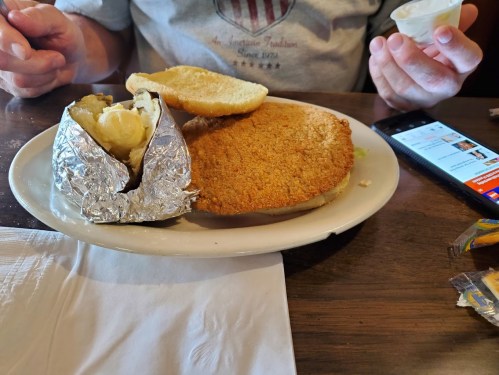 A plate with a breaded chicken breast, a foil-wrapped baked potato, and a bun, with a person holding a cup and phone nearby.