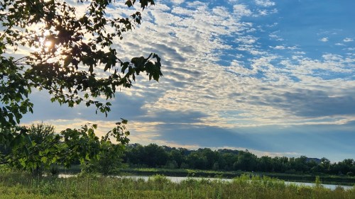 A serene landscape featuring a river, lush greenery, and a sky filled with clouds and sunlight peeking through.