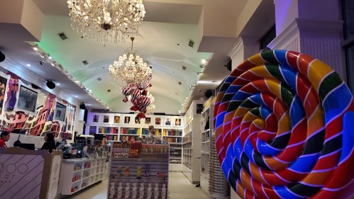 A colorful candy store interior featuring large lollipops, chandeliers, and vibrant artwork on the walls.