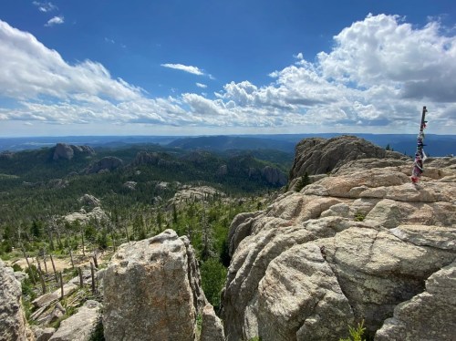 A panoramic view from a rocky summit, showcasing lush green forests and distant mountains under a partly cloudy sky.