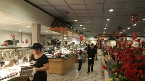 A bustling restaurant interior with a buffet, diners, and decorative flowers, creating a lively dining atmosphere.