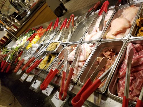 A buffet display with various raw meats, seafood, and vegetables, all arranged in metal trays with red tongs.