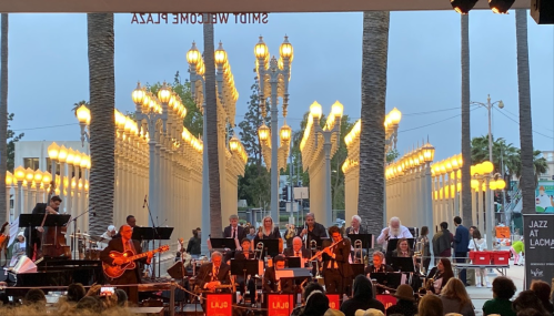 A jazz band performs outdoors at LACMA, with palm trees and illuminated street lamps in the background.