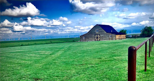 A rustic barn sits in a lush green field under a bright blue sky with fluffy clouds and distant wind turbines.