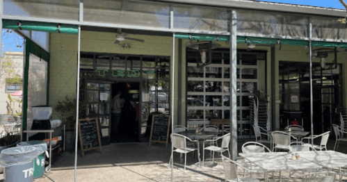 A modern café entrance with outdoor seating and large glass doors, surrounded by greenery.