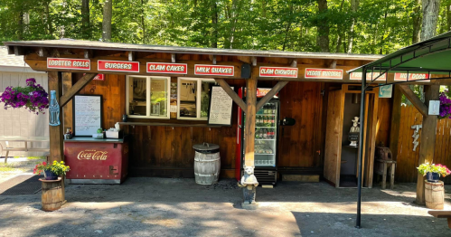 A rustic food stand in a wooded area, featuring a menu with seafood items and a Coca-Cola vending machine.