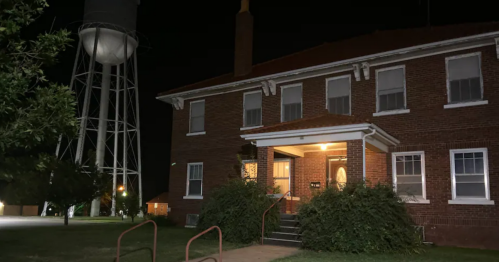 A brick building at night with a lit entrance, surrounded by greenery and a water tower in the background.