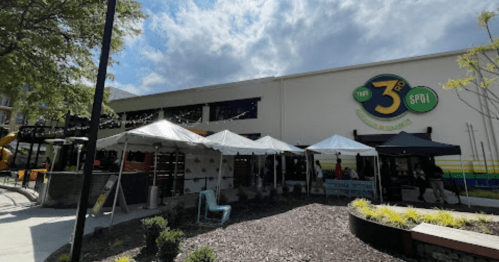 Outdoor view of The 3rd Spot, featuring tents, seating, and a colorful sign under a partly cloudy sky.