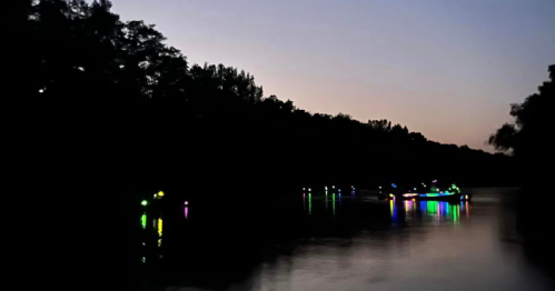 A serene river at dusk, with colorful lights reflecting on the water and silhouettes of trees in the background.