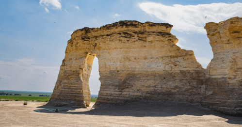 A large, natural rock formation with an arch, set against a blue sky and green landscape in the background.