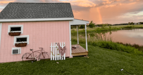 A pink house by a pond, featuring a wooden deck, bicycle, and flower baskets, with a colorful sunset in the background.