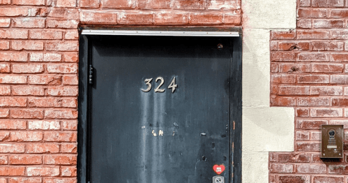 Black door with the number 324, set against a red brick wall, featuring a small intercom on the right.