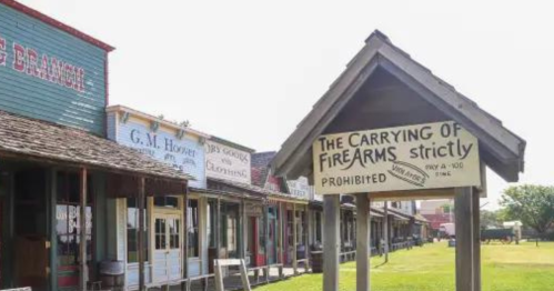 A historic street with wooden buildings and a sign stating "The Carrying of Firearms Strictly Prohibited."