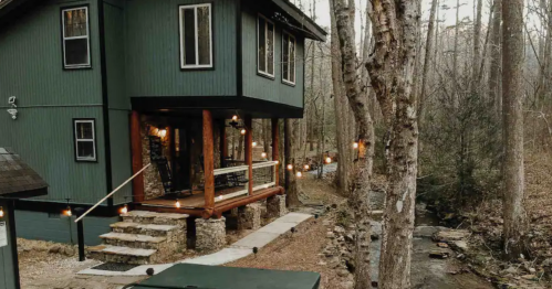 A green two-story cabin with wooden accents, surrounded by trees and a small stream, illuminated by string lights.