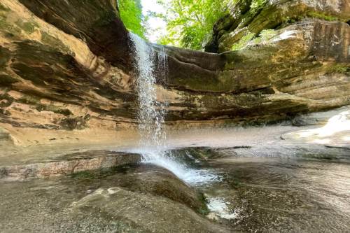 A serene waterfall cascades over rocky cliffs into a tranquil pool, surrounded by lush greenery.