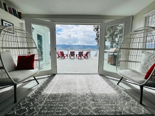 A cozy room with two hanging chairs, a patterned rug, and large doors opening to a deck with red chairs and a scenic view.