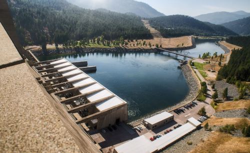 Aerial view of a dam with a river, surrounded by green hills and a clear sky.