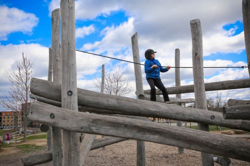 A child in a blue jacket climbs on a wooden play structure against a cloudy sky.