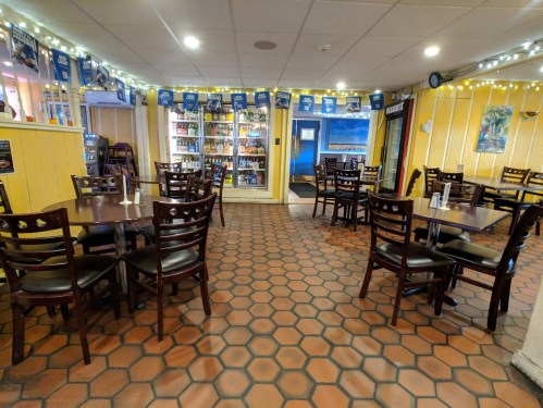 Cozy restaurant interior with wooden tables, chairs, and a colorful wall, featuring a drink fridge and decorative lights.