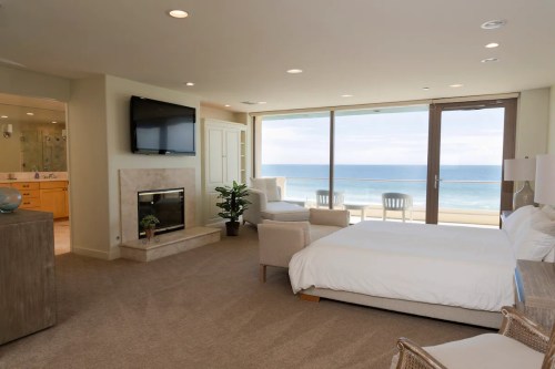 A modern hotel room with a bed, fireplace, and large windows overlooking the ocean. Bright and airy atmosphere.