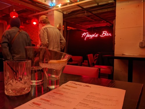 A dimly lit bar scene featuring a neon sign, drinks on the table, and patrons in the background.