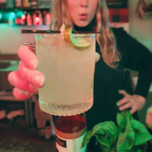 A person holding a cocktail with lime and a garnish, with a blurred background of colorful lights.
