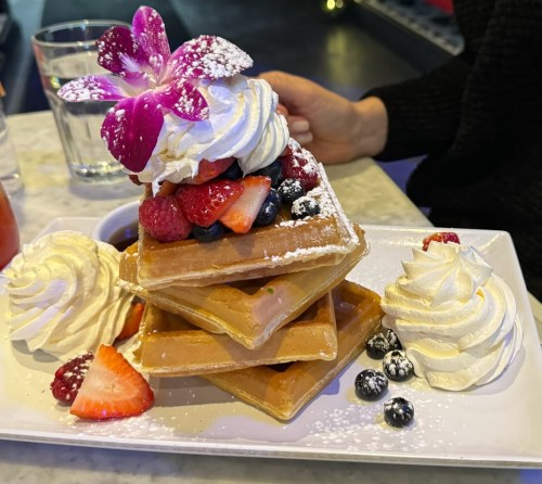A stack of waffles topped with whipped cream, berries, and a flower, served on a white plate with a side of syrup.