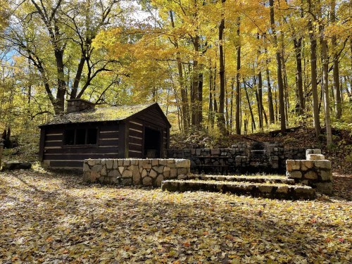 A rustic log cabin surrounded by vibrant autumn foliage and a stone structure in a serene forest setting.