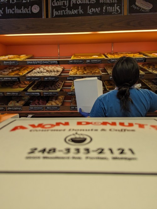 A person stands in front of a display case filled with donuts at Avon Donuts in Michigan, holding a notepad.