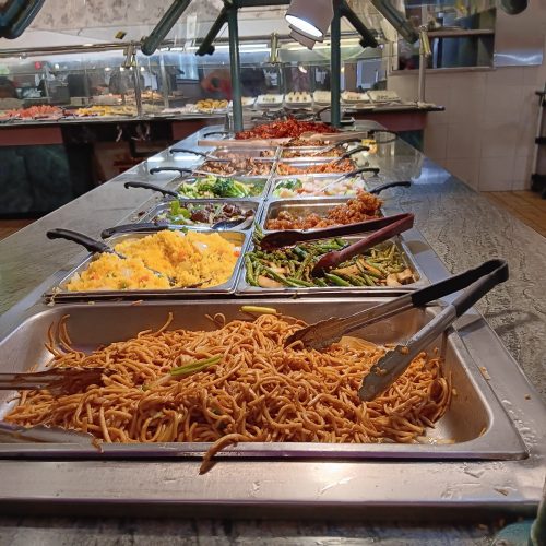 A buffet table with various dishes, including noodles, rice, vegetables, and meats, displayed in metal trays.