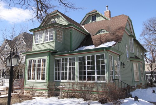 A green Victorian-style house with large windows, surrounded by snow and bare trees under a clear blue sky.