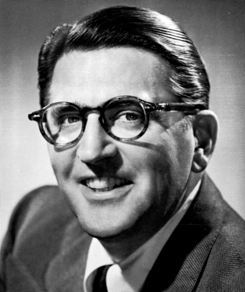 Black and white portrait of a smiling man with glasses and slicked-back hair, wearing a suit and tie.