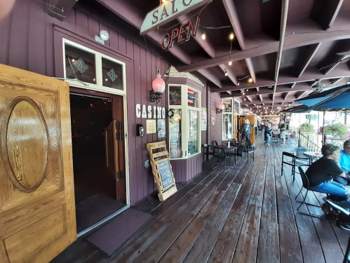 A wooden entrance to a casino with an "Open" sign, surrounded by outdoor seating and shops along a wooden walkway.