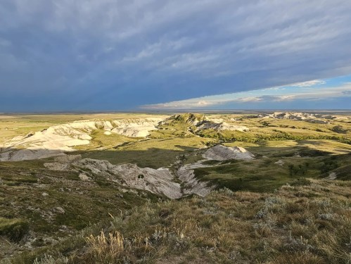 A panoramic view of rolling hills and valleys under a dramatic sky, showcasing a mix of grassland and rocky terrain.