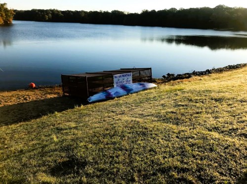 A calm lake at sunset with a kayak rental sign and a colorful kayak on the grassy shore.
