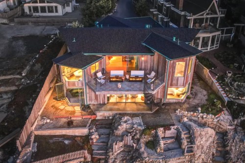 Aerial view of a modern coastal home with large windows, surrounded by rocks and a pathway, illuminated at dusk.