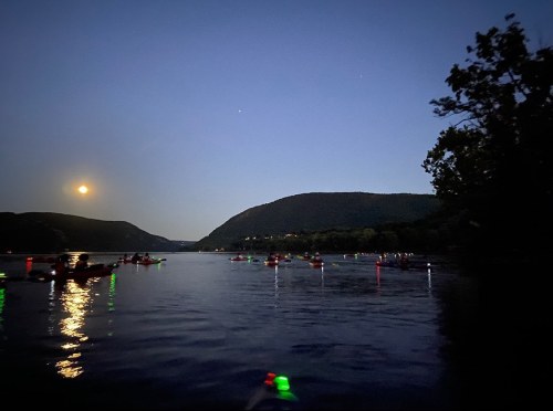 A serene night scene of a river with kayakers illuminated by colorful lights, under a full moon and mountains in the background.