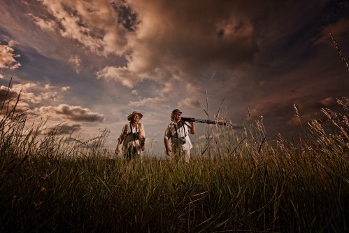 Two people walking through tall grass under a dramatic sky, one carrying a camera on a tripod.