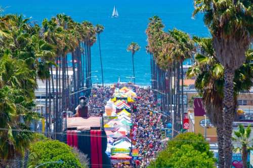 A vibrant street fair lined with palm trees, colorful tents, and a view of the ocean in the background.