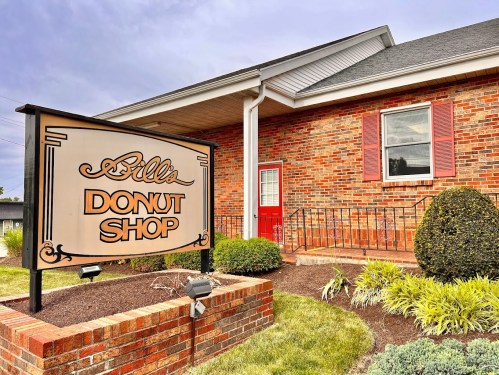 A brick building with a sign reading "Silla Donut Shop" and a red door, surrounded by green landscaping.