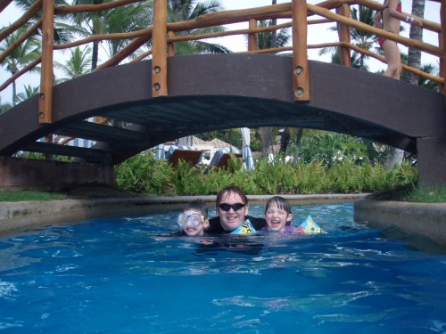 A man and two children smile while swimming under a wooden bridge in a blue pool surrounded by greenery.